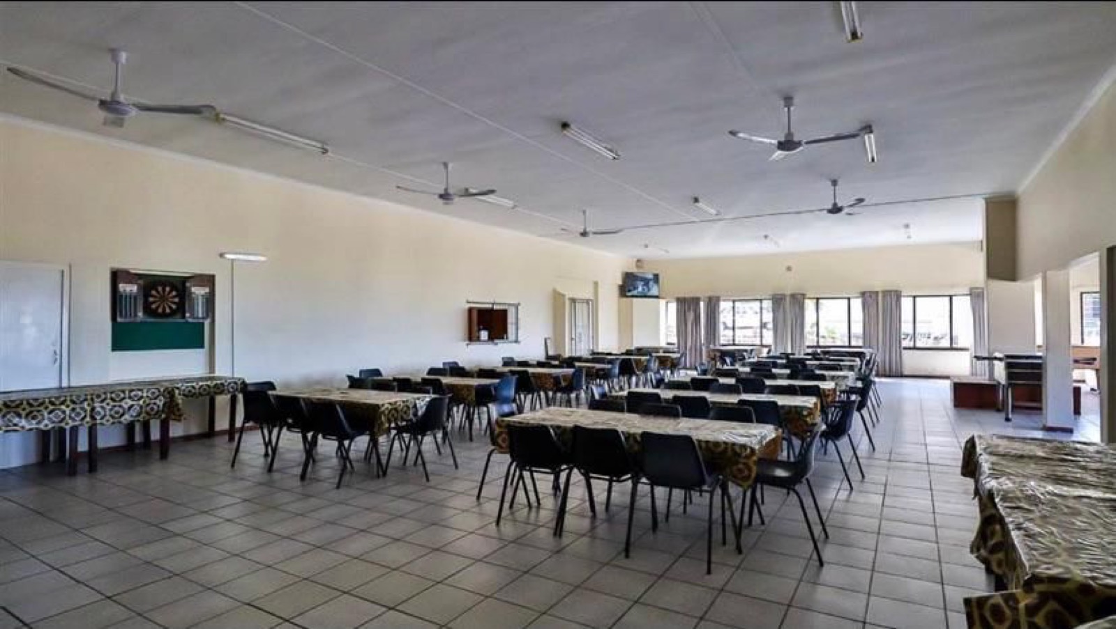 Tetelestai Recovery Centre indoor recreation and dining lounge with tables and chairs, a dartboard, wall-mounted TV, ceiling fans, and large windows